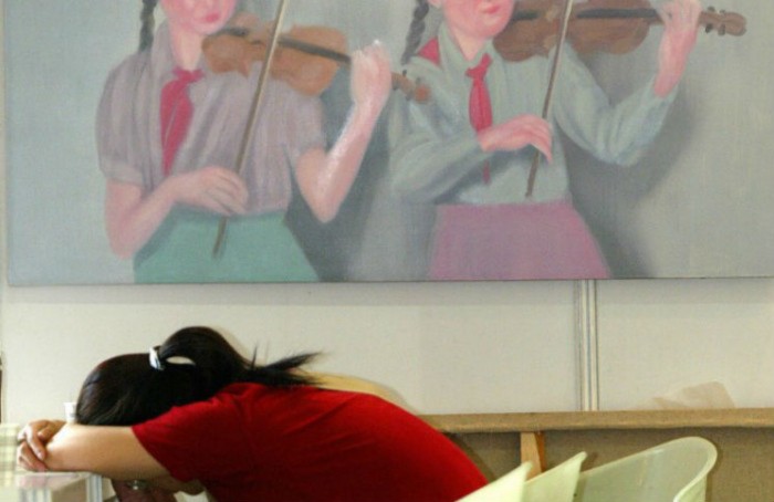 Một người phụ nữ tranh thủ ngủ gục trên bàn ngay cạnh một bức tranh sơn dầu vẽ hai đoàn viên thanh niên chơi violin tại Phòng trưng bày Nghệ thuật Mùa xuân Thượng Hải, ngày 3/6/2004. (LIU JIN/AFP/Getty Images)